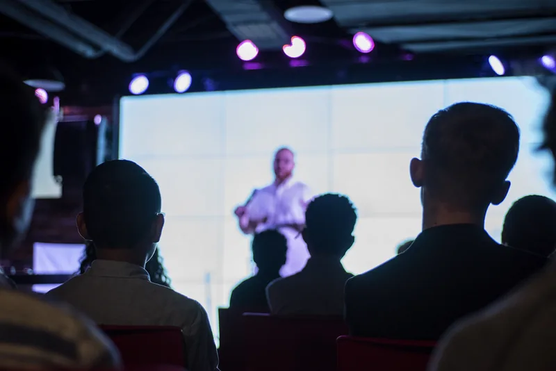 An audience watching a speaker on a bright screen in a dark room