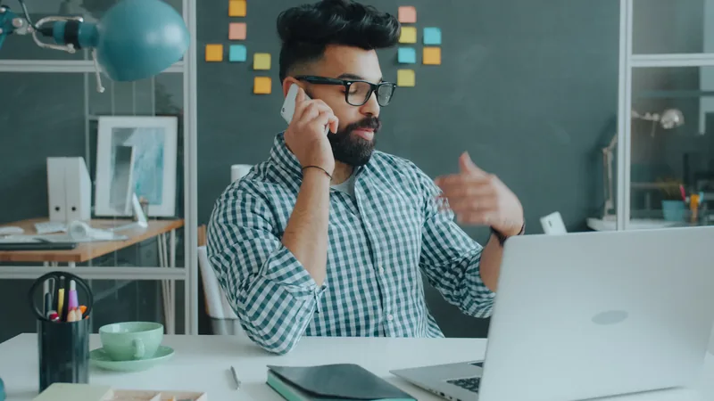 A man with a beard and glasses talking on a phone in a workspace