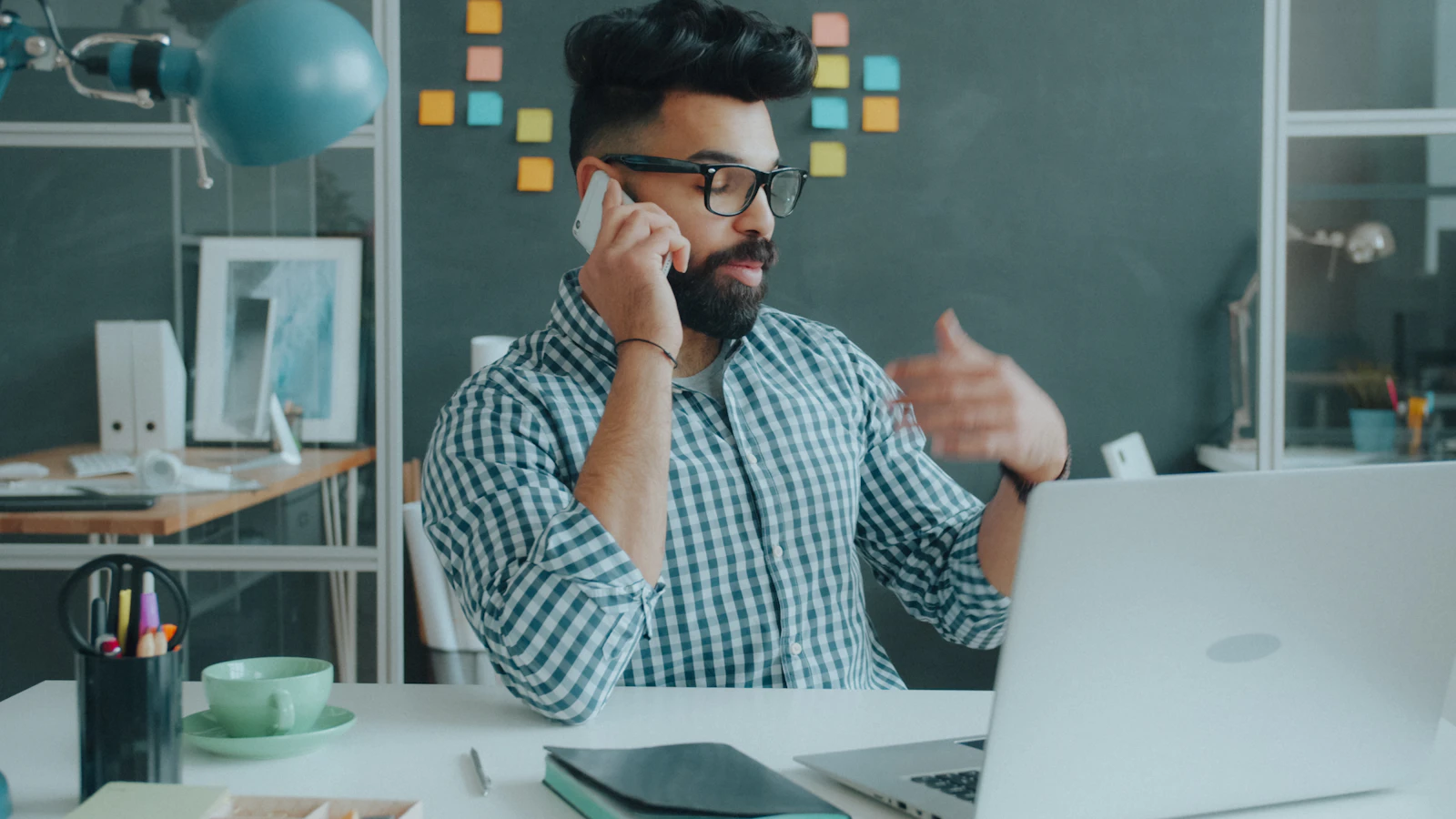 A man with a beard and glasses talking on a phone in a workspace